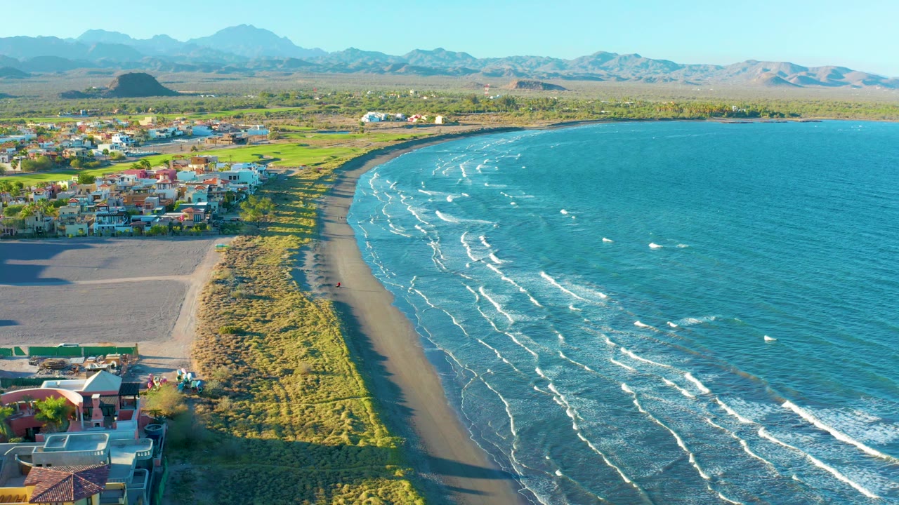 Curving sandy beach with surf lines, dunes, town buildings, and mountains.