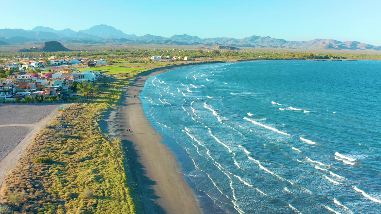 Aerial view over Loreto, Baja California Sur, showing a curved dark beach beside blue water, grassy dunes, low buildings, distant mountains, and a tiny walker near the lower shoreline.