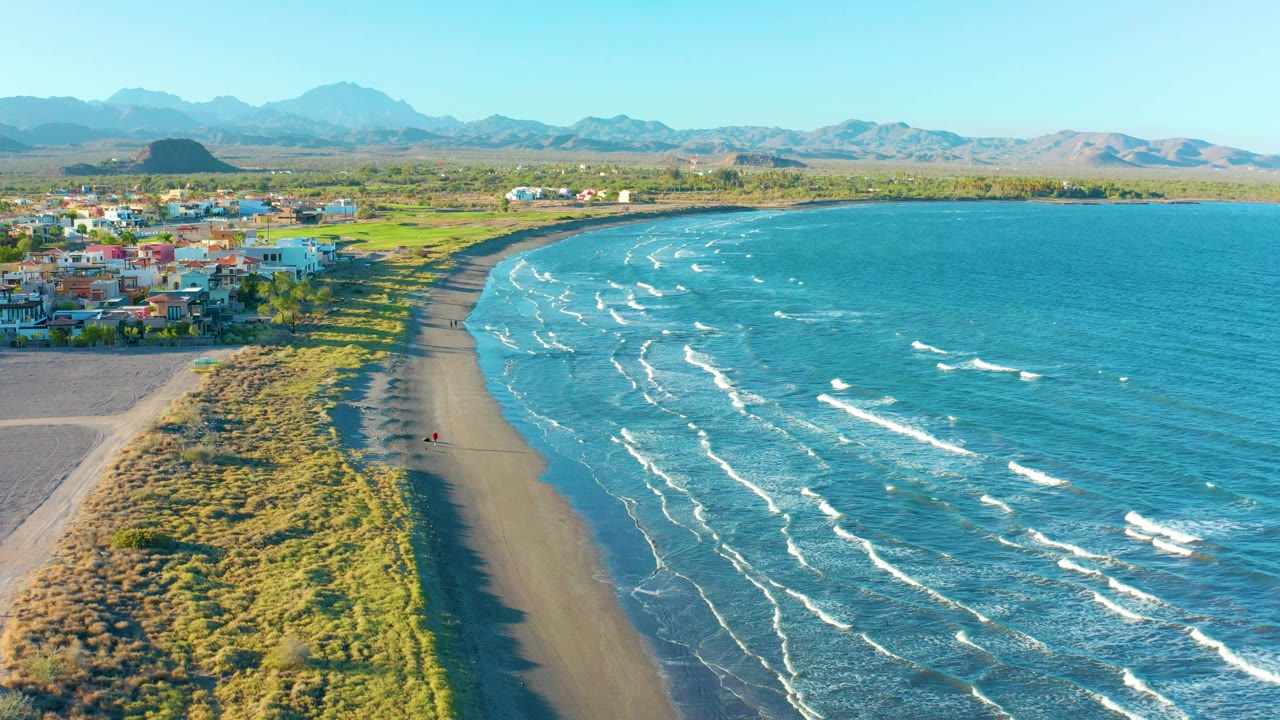 Drone view along a curved beach with surf, dunes, buildings, mountains, and a small person on the sand.