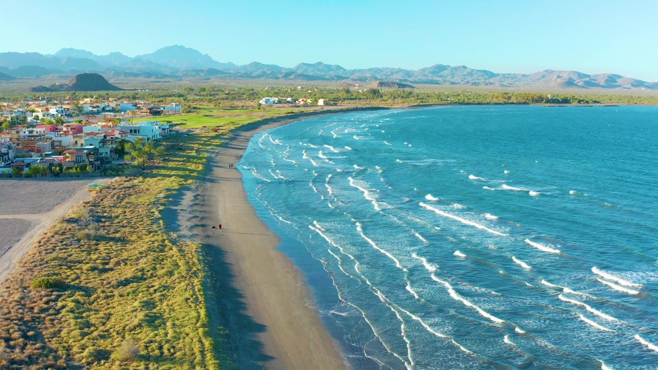 Aerial frame of Loreto's curved shoreline, with turquoise-blue water filling the right side, narrow white surf lines, dunes along the beach, low buildings at left, and a tiny person on the sand.