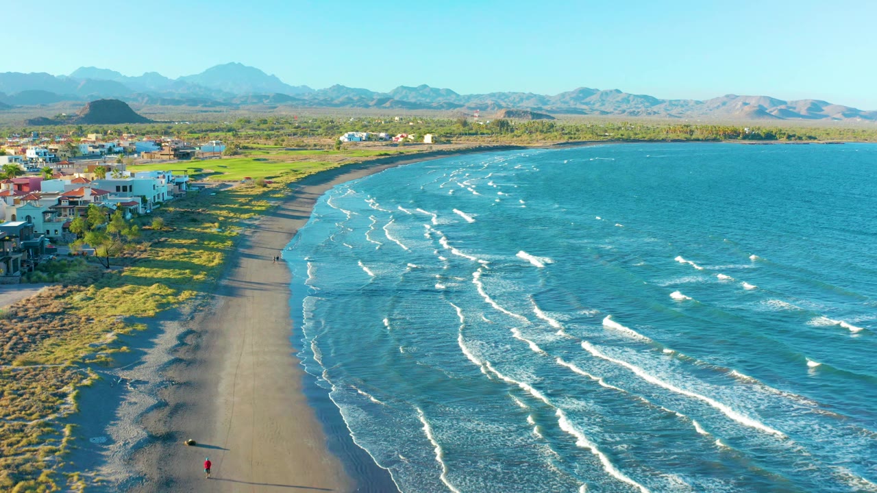 Curving beach with white surf lines, houses, grass, mountains, and a small walker on the sand.