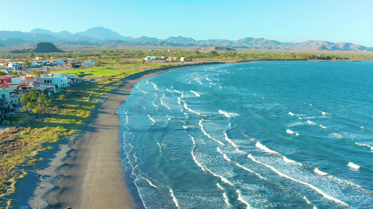Aerial frame over Loreto, Baja California Sur, Mexico, with more ocean filling the right side, the beach curving past grassy shore and houses at left, distant mountains across the horizon, and no clearly visible people.