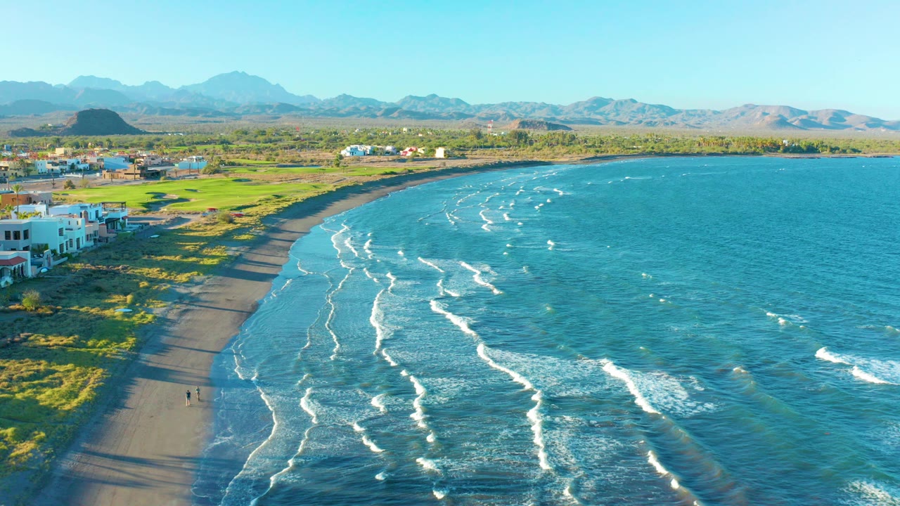 Aerial frame over Loreto, Baja California Sur shows a curved sandy beach with white wave lines, low buildings at left, green coastal ground, distant mountains, and tiny people on the sand.