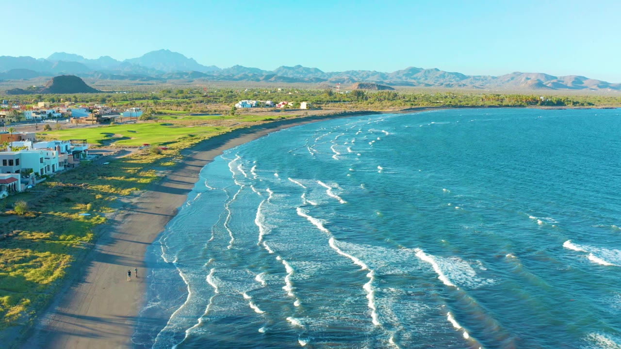 Drone view along a sandy shoreline with surf and town-edge buildings.