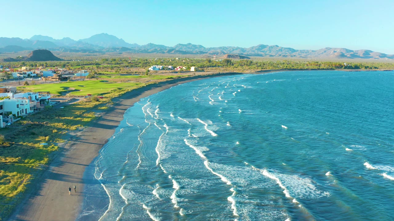 The Loreto coastline bends across the frame with white surf marks spread through the blue water, green land and low buildings inland, and a broad mountain horizon beyond.
