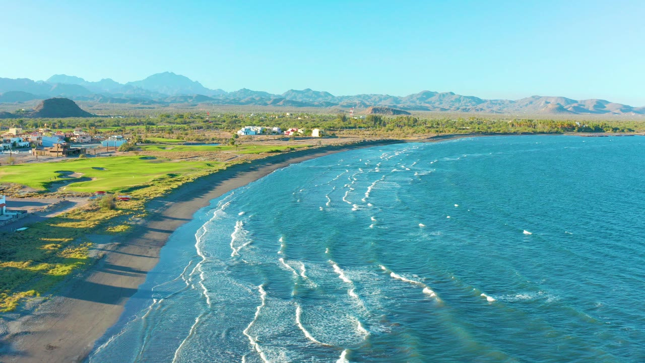 Aerial frame over Loreto, Baja California Sur shows blue-green water with white wave lines meeting a dark sand beach, green fields and buildings at left, distant mountains, and no visible people.