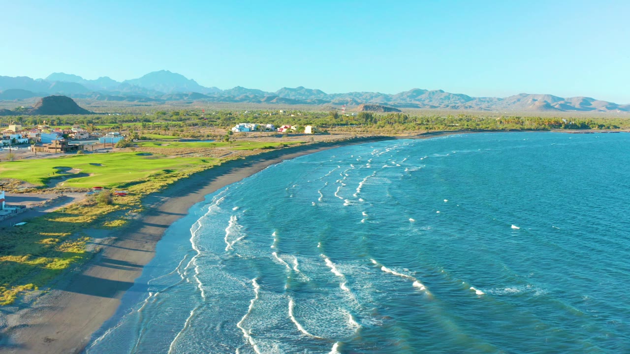 Aerial view in Loreto, Baja California Sur shows the curved dark sand shoreline slightly farther along, with surf streaks across the water, green field patches inland, low buildings, mountains, and no visible people.