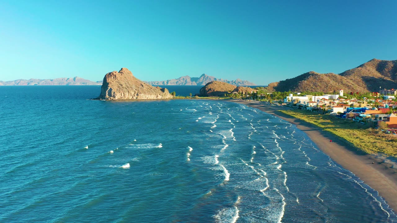 Aerial view over Loreto, Baja California Sur, with blue ocean in the foreground, white wave lines approaching a sandy beach, rocky hills, distant mountains, low buildings, and no visible people.
