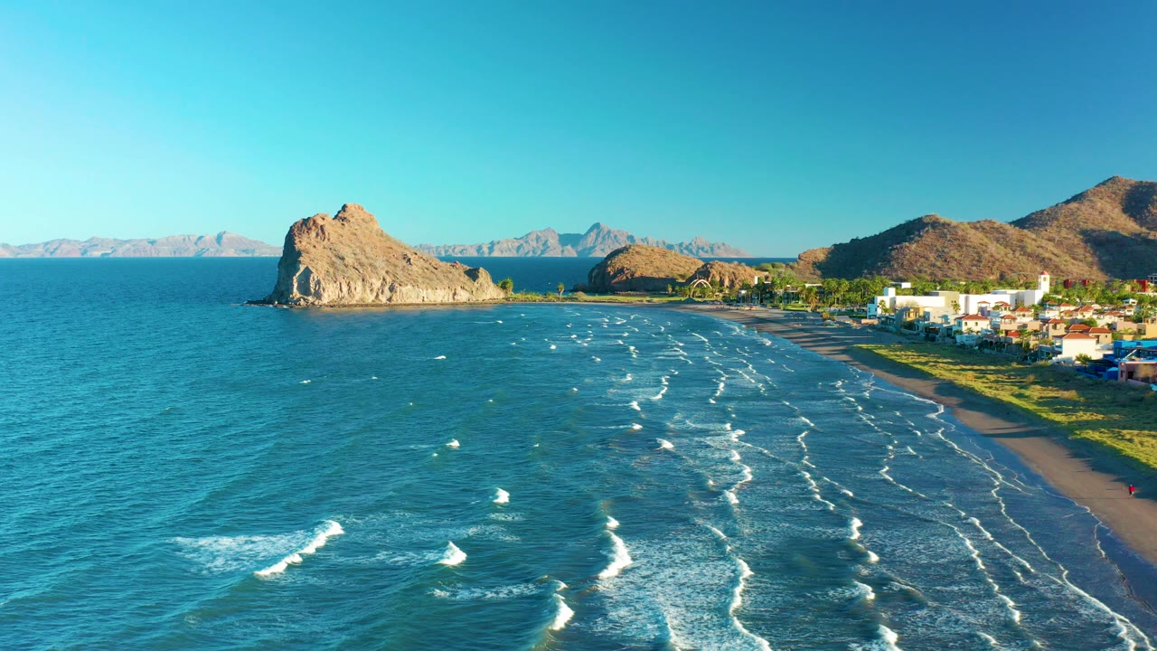 Opening aerial frame over Loreto, Baja California Sur, with blue water, white surf lines, a sandy beach, shoreline buildings, rocky hills, distant mountains, and no visible people.