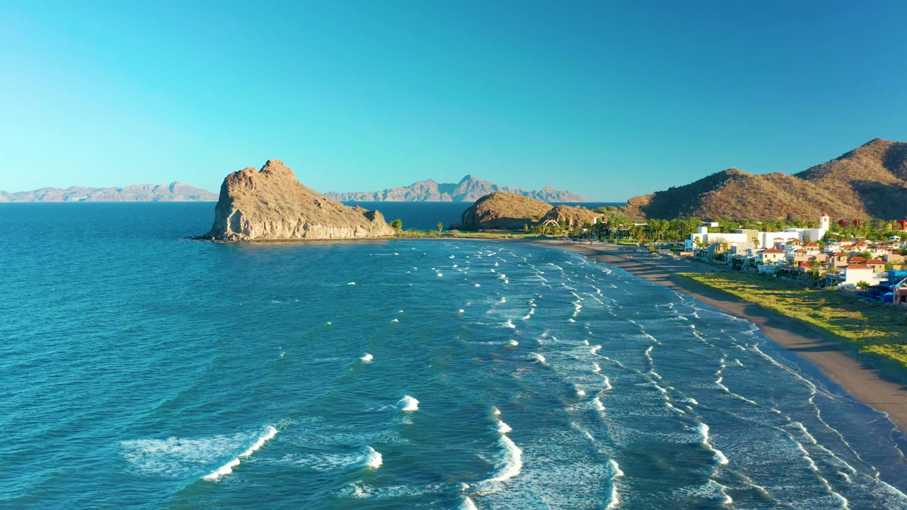 Loreto coastal aerial with the rocky outcrop left of center, rows of white waves crossing the bay, shoreline buildings on the right edge, distant mountains on the horizon, and no visible people.