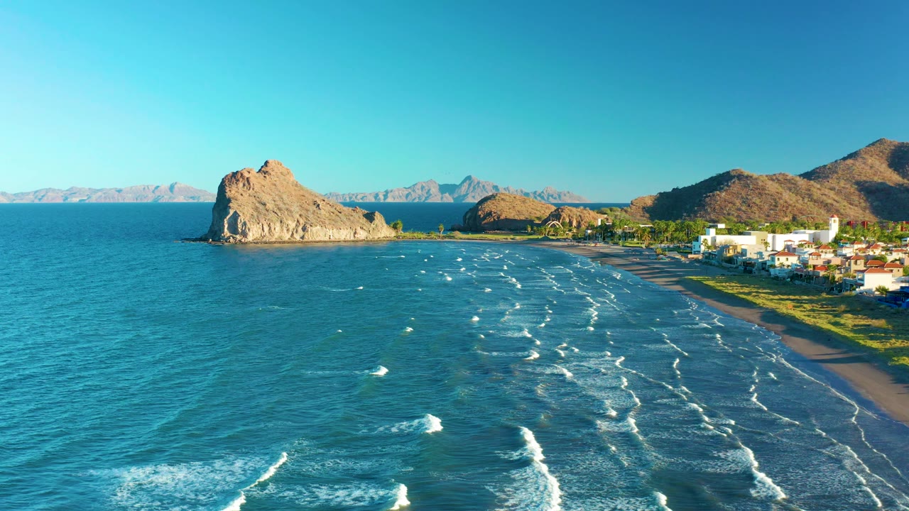 Aerial shoreline view with open water, surf, beach, rocky point, and buildings.