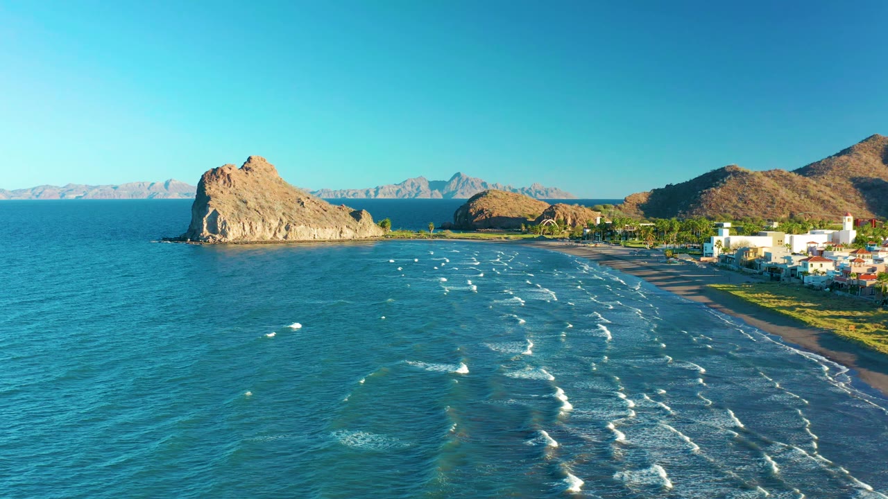 Aerial frame from Loreto, Baja California Sur, Mexico, showing the surf lines slightly farther across the bay, the offshore tan rock near the left center, white buildings on the right edge, distant mountains, and no visible people.
