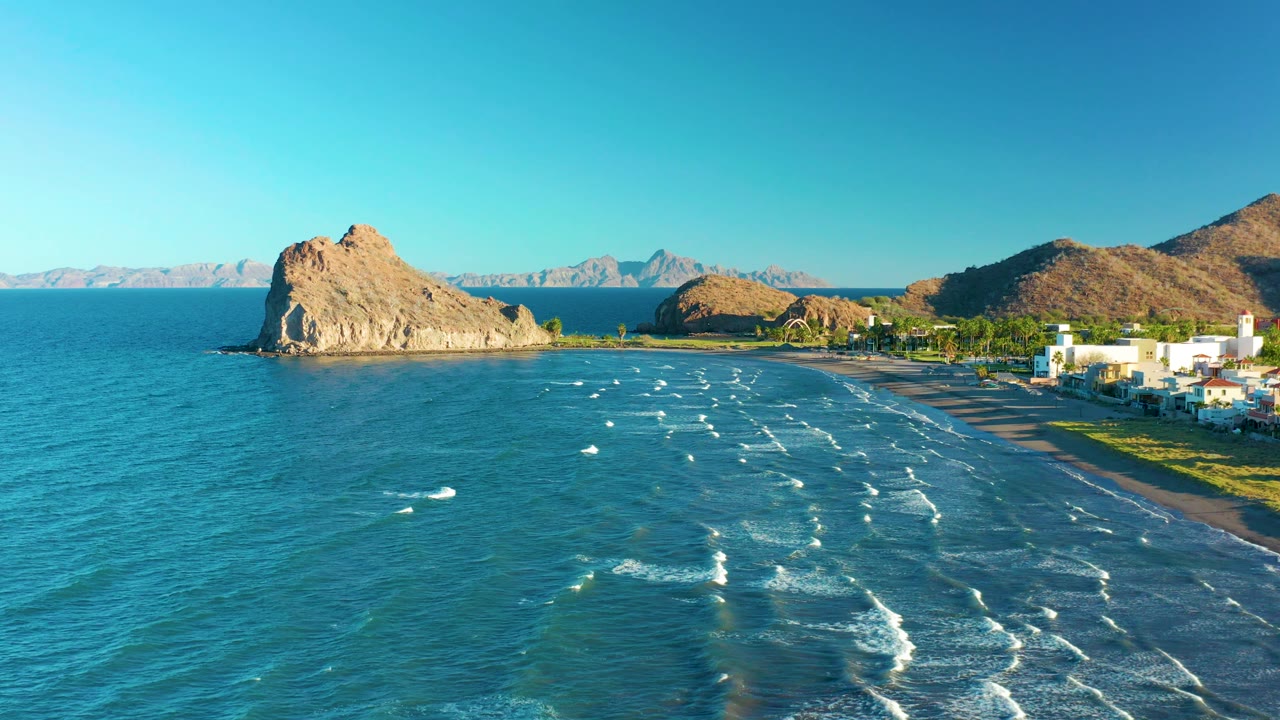Aerial frame from Loreto, Baja California Sur, Mexico, showing a broad curve of beach, scattered white surf marks across blue-green water, the tan rock formation at left, coastal buildings at right, mountain slopes behind, and no visible people.