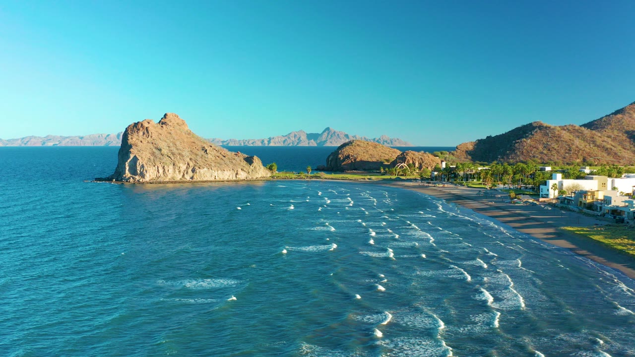 Wide aerial frame of Loreto, Baja California Sur, Mexico, with turquoise water, white wave lines, a sandy beach, a tan rock formation near shore, distant mountains, coastal buildings at right, and no visible people.