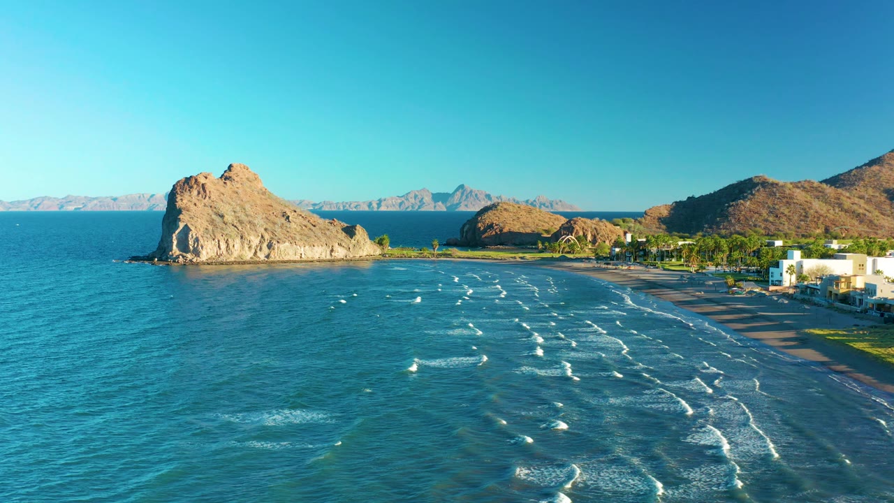 Loreto coastal aerial frame at 0:02 showing deeper blue water at left, shallow wave bands in the center, the sandy beach curving along the right, a tan rock formation near shore, distant mountains, and no visible people.