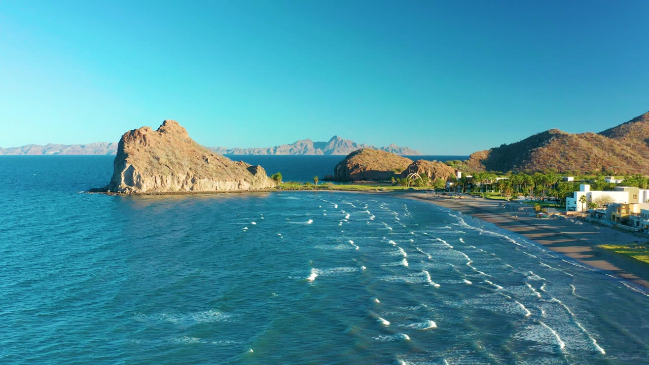 Drone-like view of ocean waves, a beach, coastal buildings, and a rocky headland.