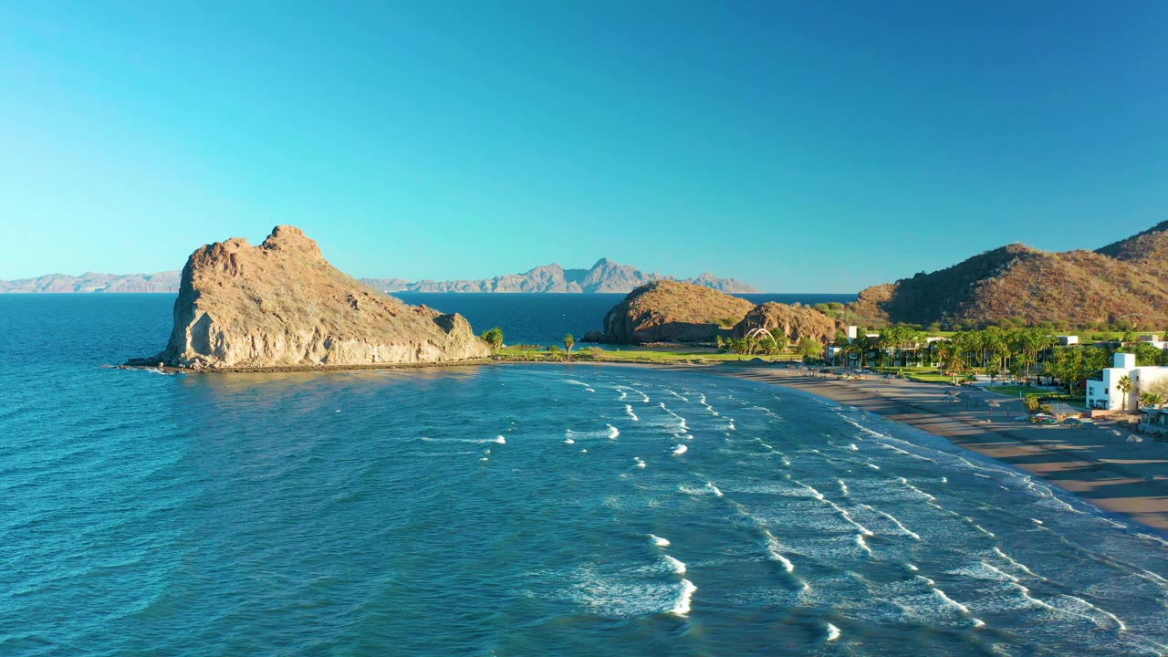 Aerial frame from Loreto, Baja California Sur, showing a large tan rocky outcrop left of center, angled white wave lines across blue-green water, beach to the right, hills on the horizon, and no visible people.