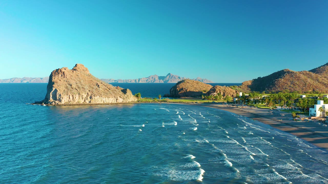 High aerial view in Loreto, Baja California Sur, with the rocky outcrop held near the left side, shallow turquoise water and foamy surf in the foreground, beach and palms to the right, and no visible people.