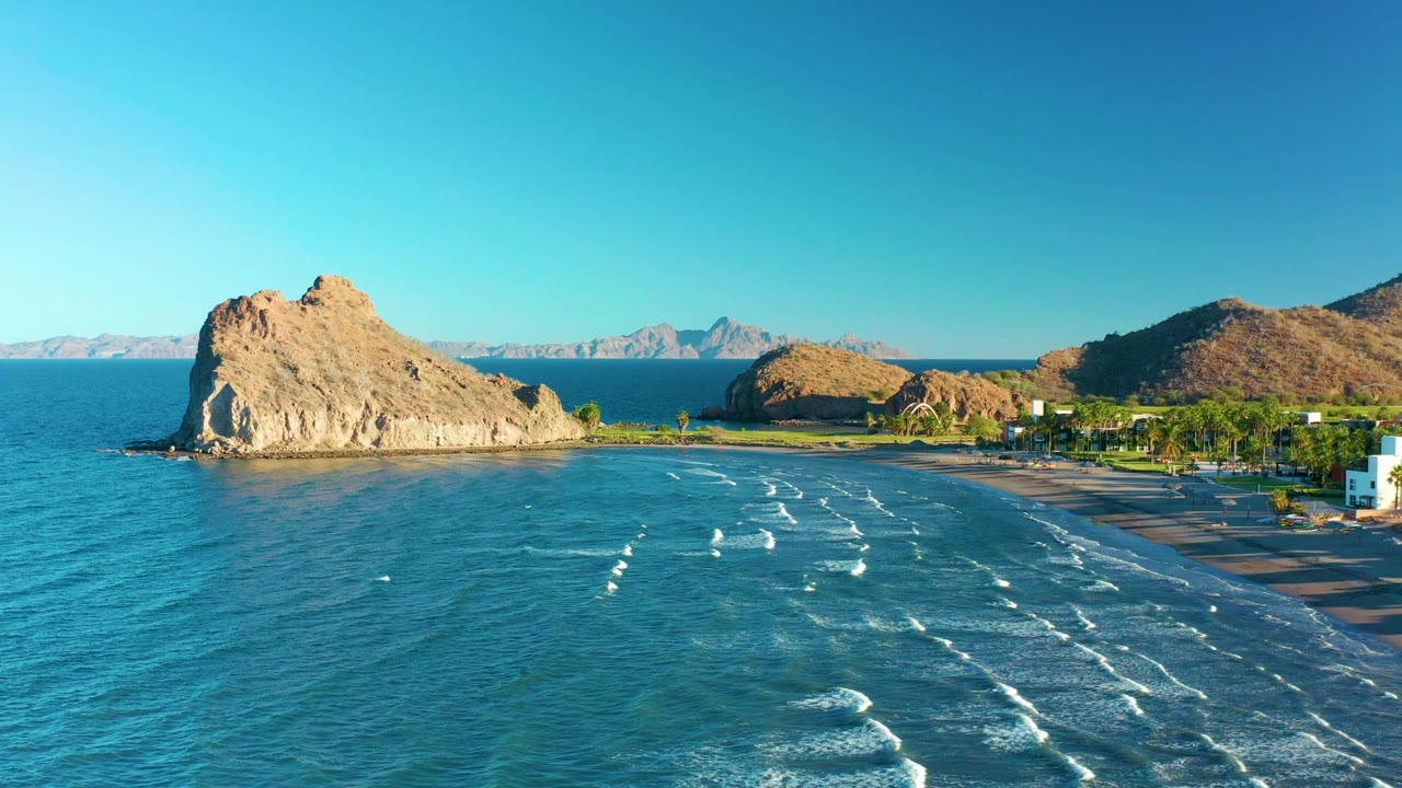 Aerial frame in Loreto, Baja California Sur, where the tan rock formation sits near the left edge, broken white surf spreads across the water, the beach runs along the right side, and no visible people appear.