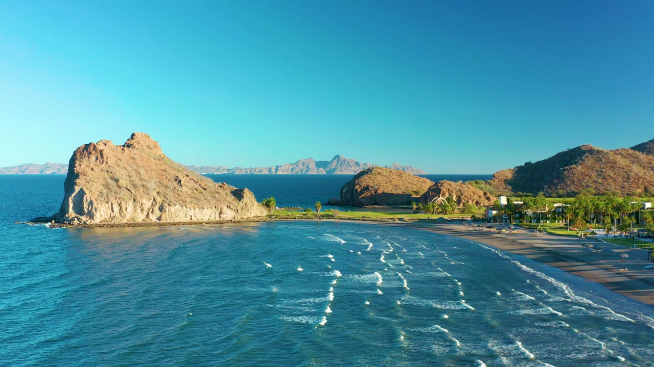 Aerial view in Loreto, Baja California Sur, Mexico, showing turquoise water, white wave lines, a tan rocky outcrop, sandy beach, distant mountains, and no visible people.