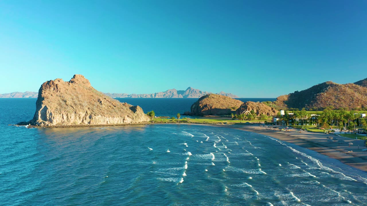 Aerial coastal frame from Loreto, Baja California Sur, Mexico, with the rocky outcrop on the left, wave foam spread across the bay, beach and palms on the right, and no visible people.