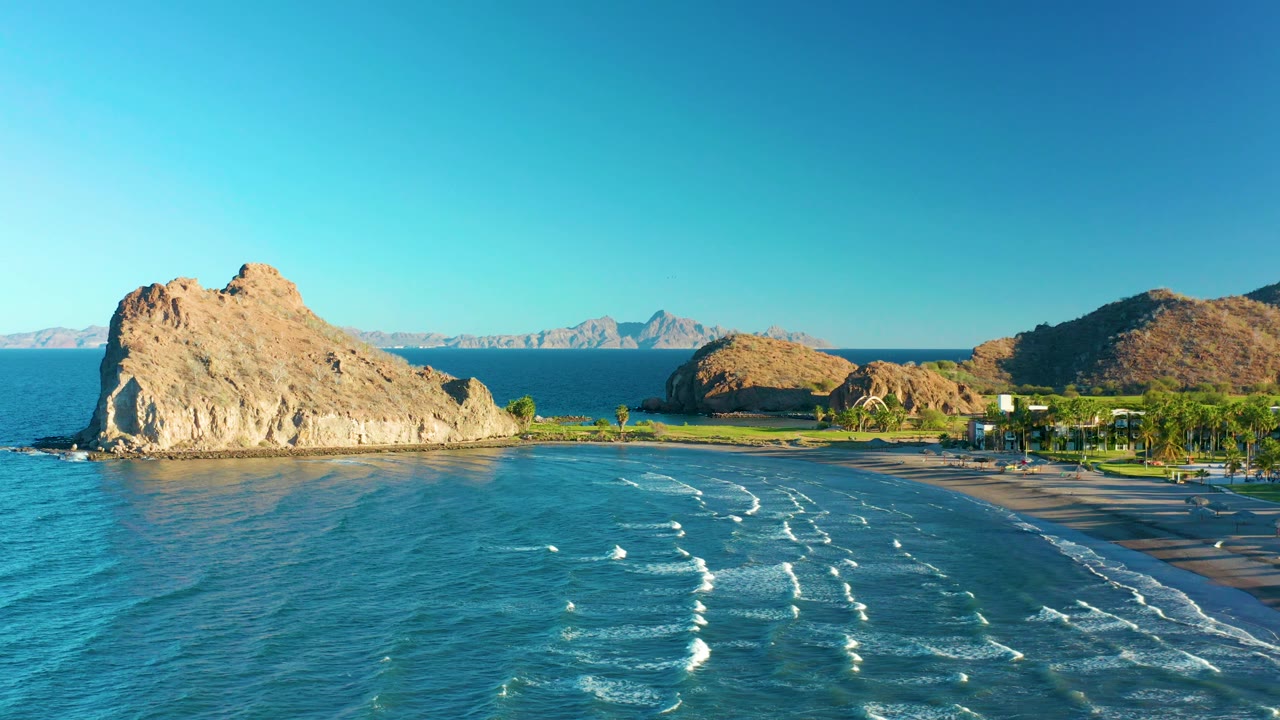 Wide aerial of ocean waves rolling toward a beach below rocky coastal hills.