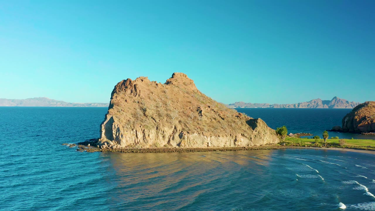 Opening aerial view of a tan rocky outcrop in Loreto, Baja California Sur, with blue water in front, a sandy shore to the right, distant landforms on the horizon, and no visible people.