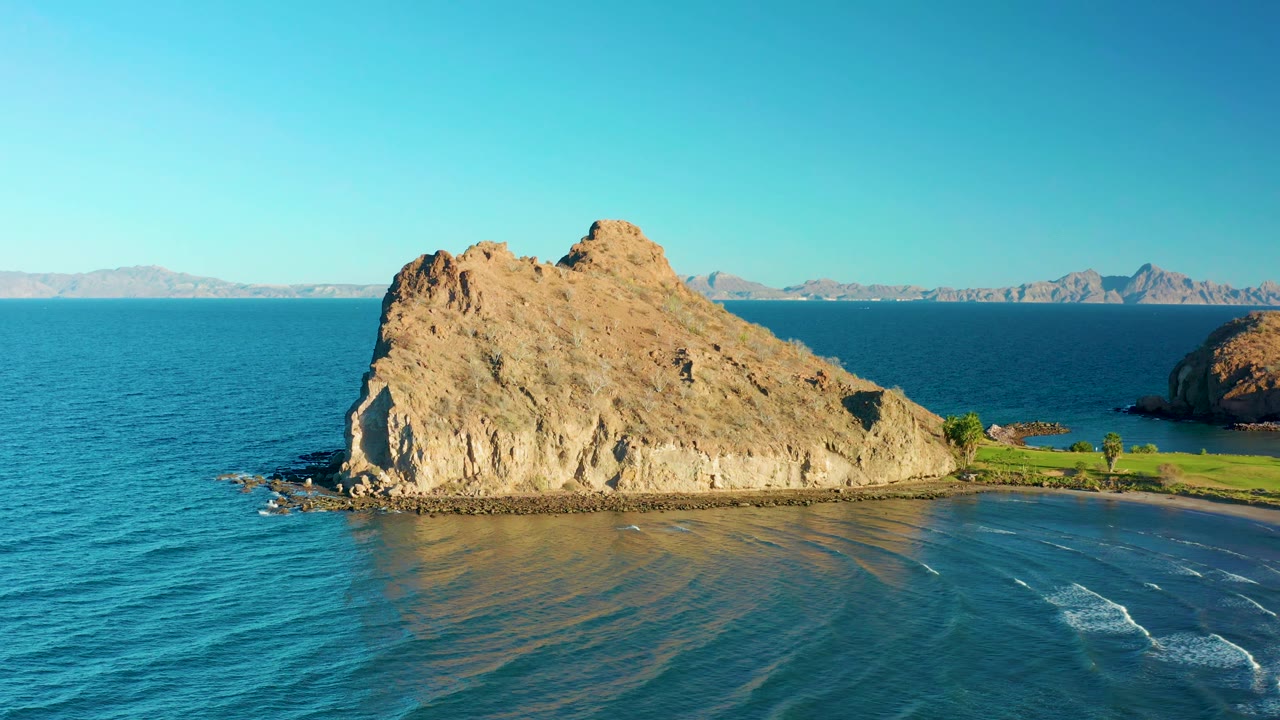 Aerial-style frame of the Loreto rocky outcrop with rippled blue water in the foreground, a thin beach curve, green grass to the right, and no visible people.