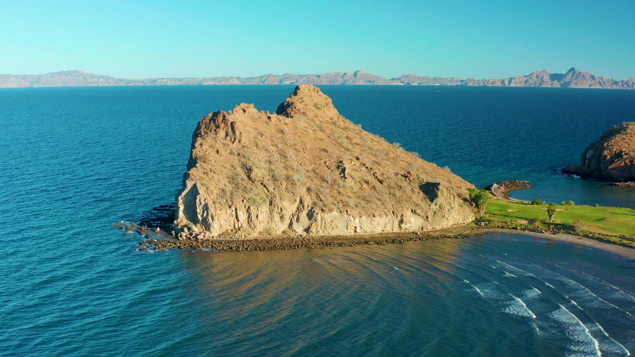 At 0:00, an aerial view in Loreto, Baja California Sur shows a tan rocky outcrop, blue ocean, shallow beach water, green shoreline grass, distant mountains, and no visible people.
