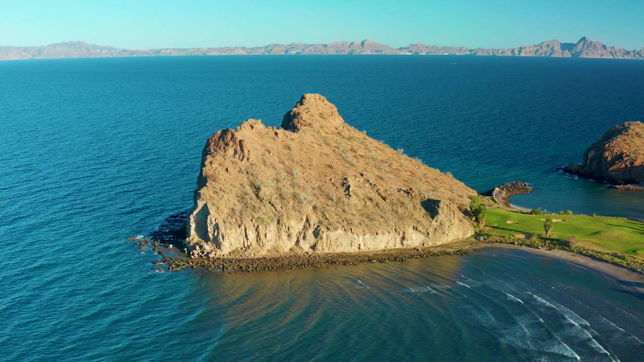 Aerial view near Loreto, Baja California Sur showing a tan rock formation, blue water, a small beach edge, distant mountains, and no visible people at the start of the clip.