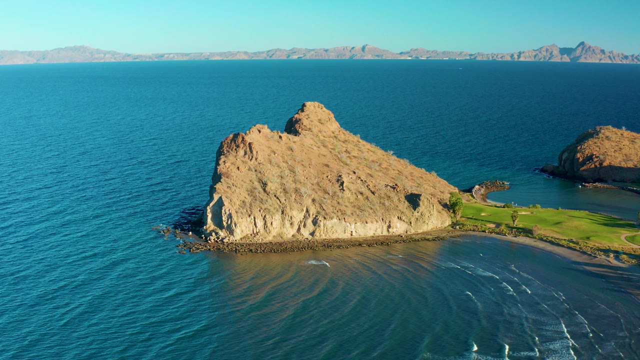 Opening frame of Loreto, Baja California Sur, with a tan rock formation extending into blue water, a narrow beach and green shore at right, distant mountains on the horizon, and no visible people.