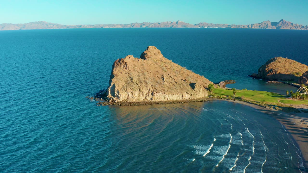 Aerial view in Loreto, Baja California Sur, Mexico, showing a tan coastal rock formation beside blue ocean, a sandy beach, distant mountains, clear sky, and no visible people.