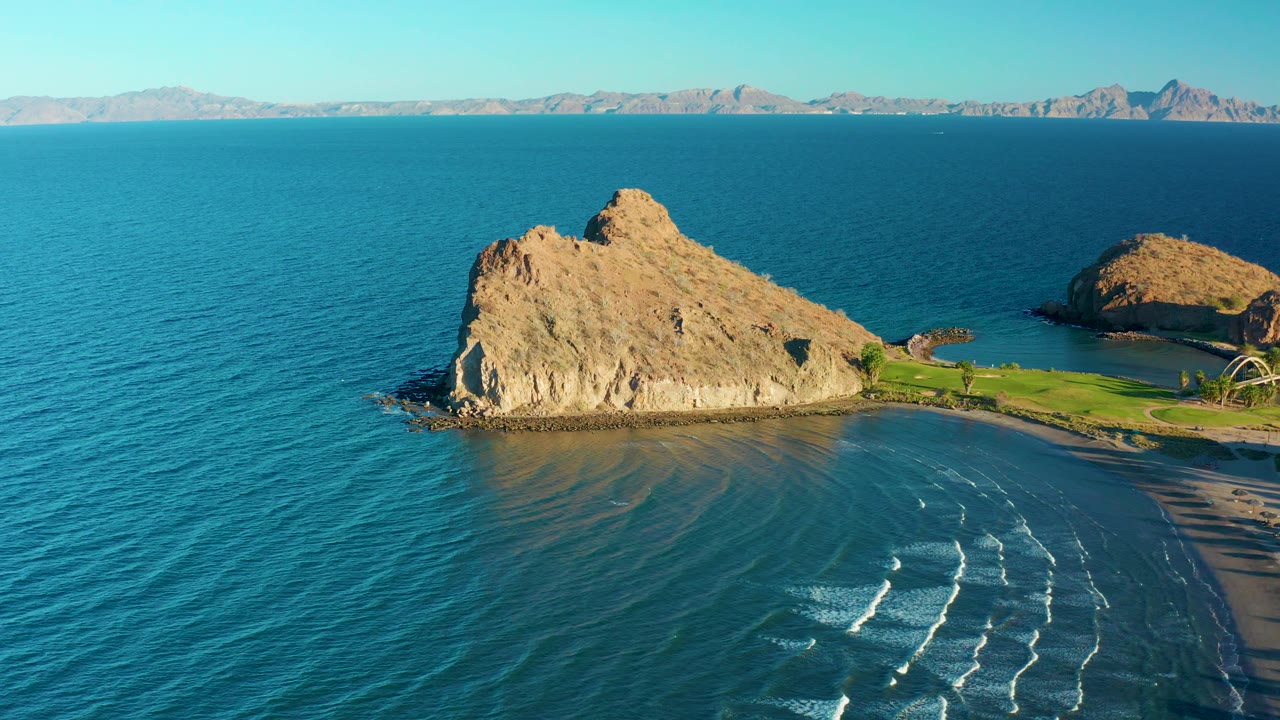 The Loreto aerial view holds on the tan rock, blue-green bay water, sandy shoreline, repeating white surf bands, distant mountain horizon, and no visible people.