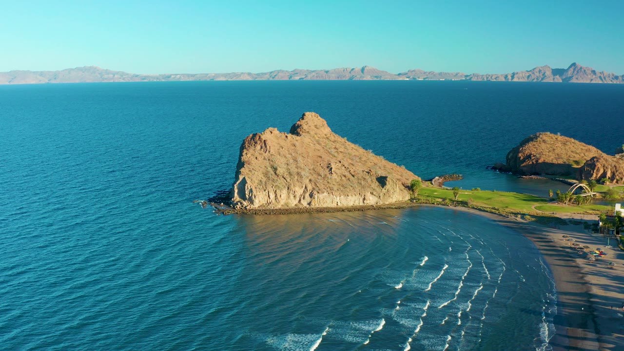 Opening frame over Loreto, Baja California Sur, Mexico, showing blue ocean around a tan rocky outcrop, a curved sandy beach, green shore area, distant mountains, and no visible people.