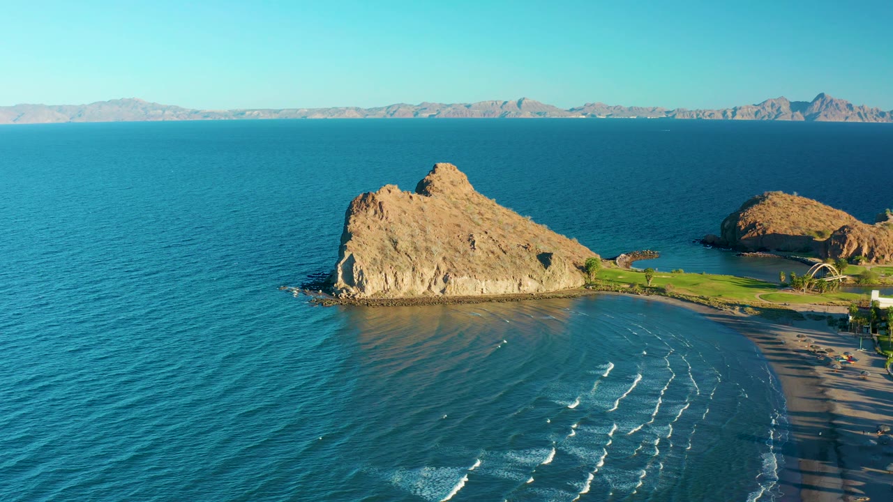 At 0:02, the Loreto coastline remains in a wide aerial composition, with the tan rock outcrop prominent, calm blue water surrounding it, beach waves below, and no visible people.