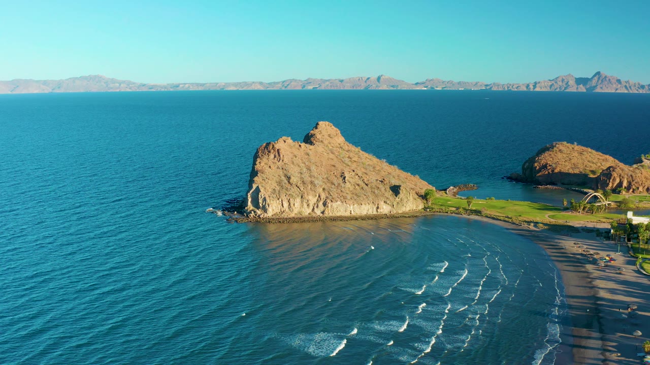 At 0:03 in Loreto, Baja California Sur, Mexico, the frame shows a broad sweep of blue ocean left of the rocky outcrop, small surf lines on the beach, and no visible people.