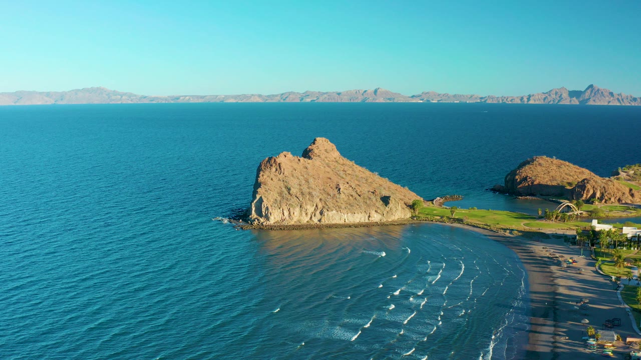 Opening aerial frame over Loreto, Baja California Sur, showing a tan rocky outcrop beside a curved sandy beach, blue-green water, distant mountains, clear sky, and no visible people.