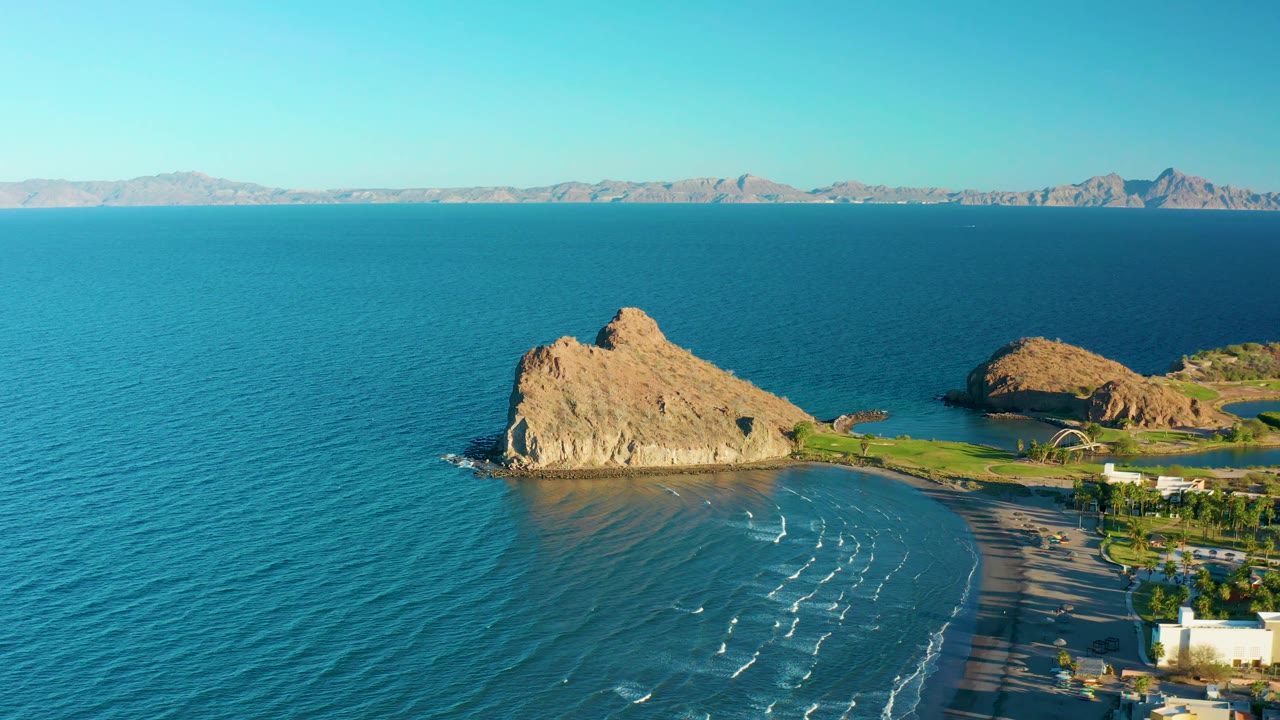 Second frame over Loreto shows the rocky outcrop slightly more centered above the beach, with white wave streaks, low buildings at lower right, distant mountains, and no visible people.