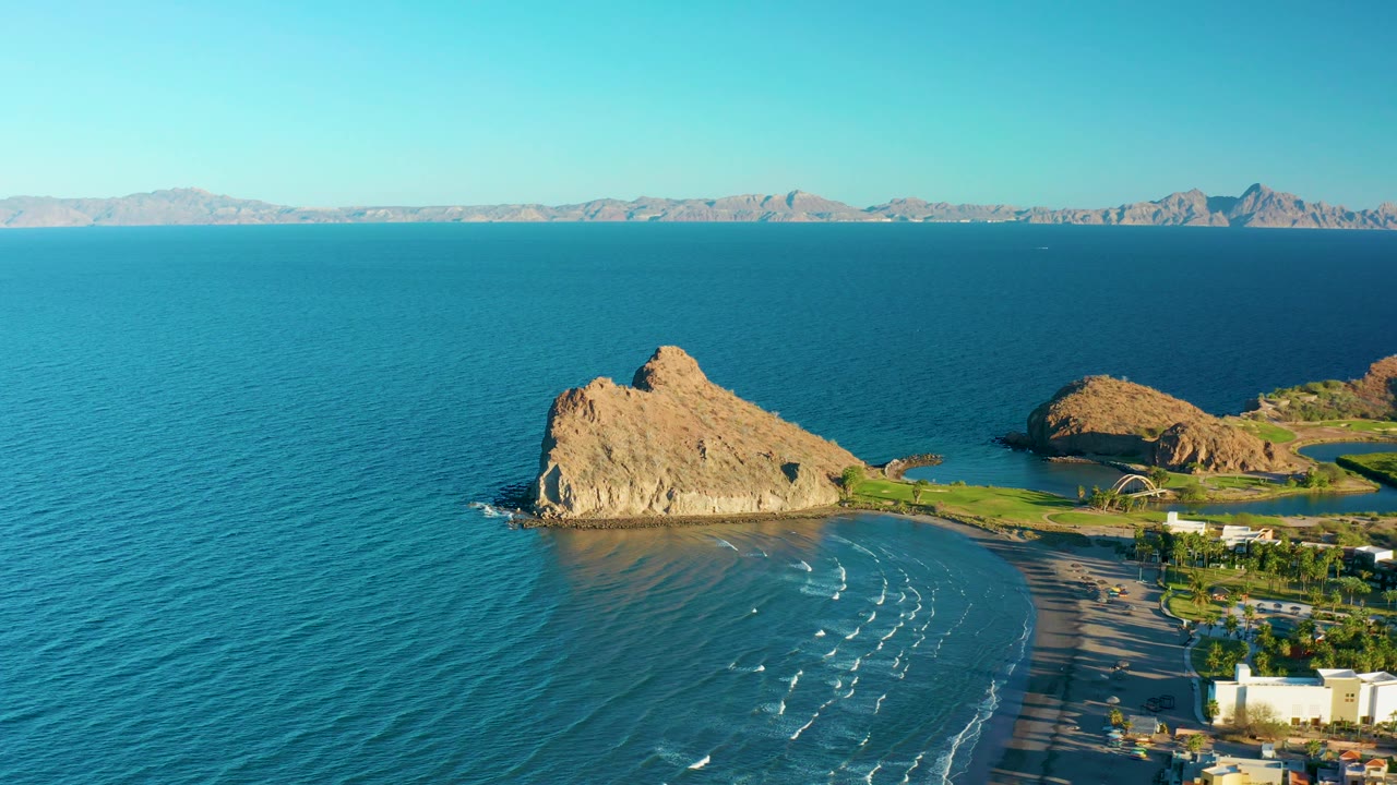 At 0:03, the Loreto coastline remains in a wide aerial composition with the tan rock, shallow surf bands on the beach, distant mountain ridge, and no visible people.
