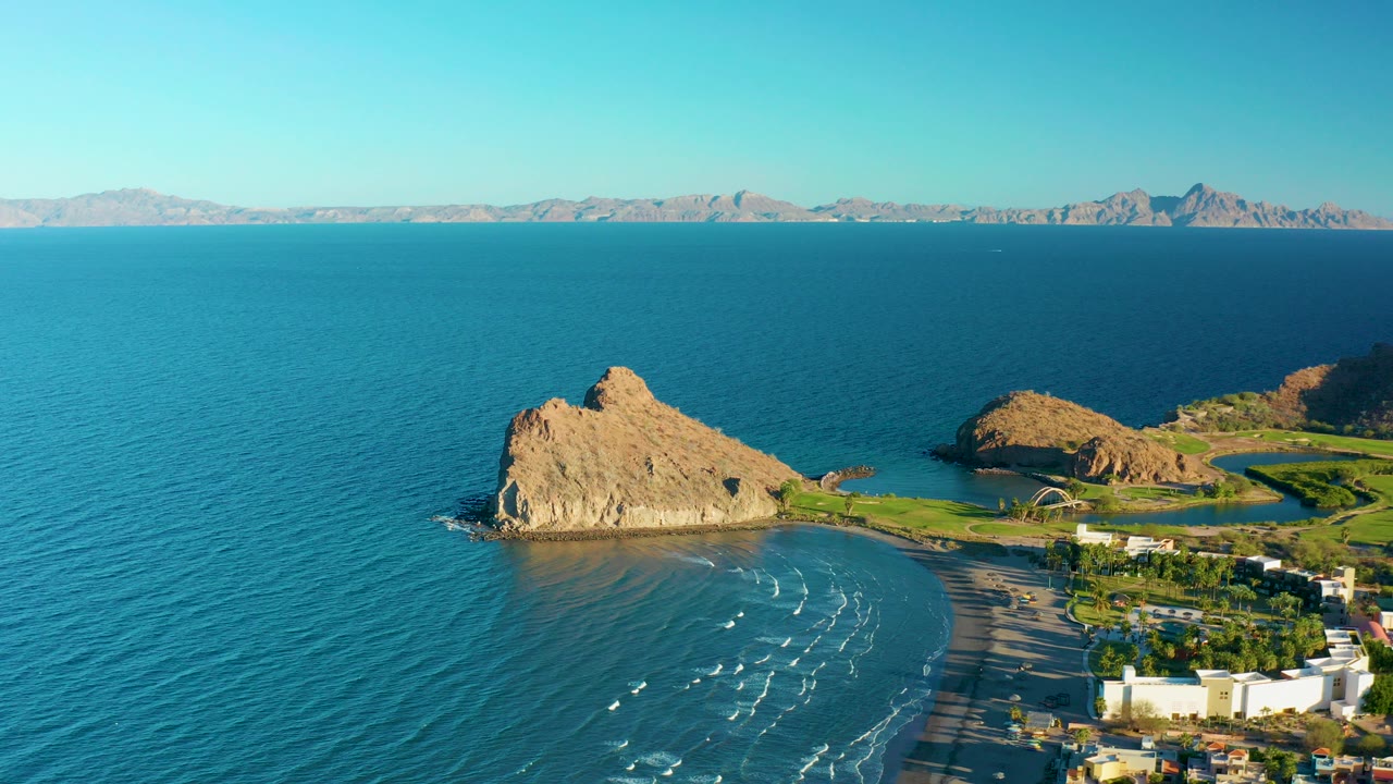 Aerial frame over Loreto, Baja California Sur showing a tan rocky outcrop, blue ocean, curved sandy beach, distant mountains, clear sky, and no visible people.