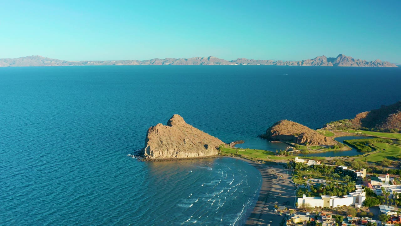 At 0:00, an aerial view over Loreto, Baja California Sur shows turquoise ocean around a tan rocky point, a sandy beach, shoreline buildings, distant mountains, clear sky, and no visible people.