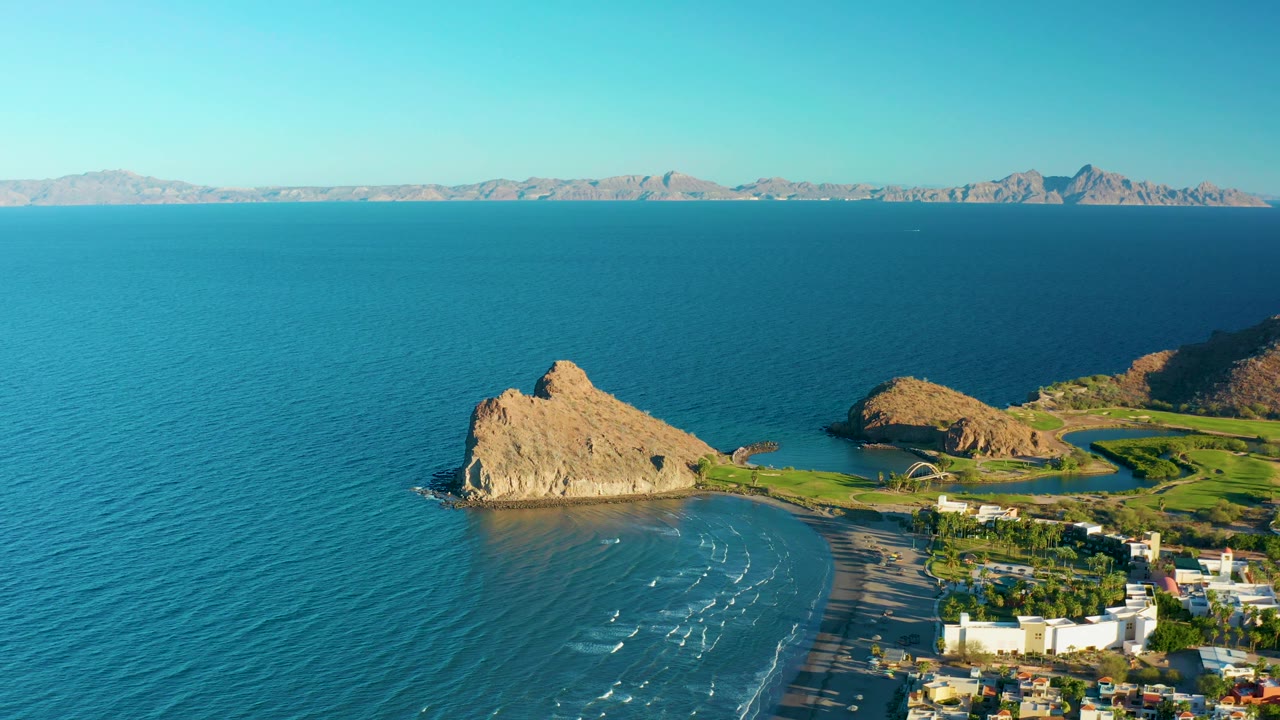 At 0:02, the frame shows a wide Loreto coastline view with calm turquoise water, the tan rock point projecting into the sea, pale beach surf, white shoreline structures, mountain backdrop, and no visible people.