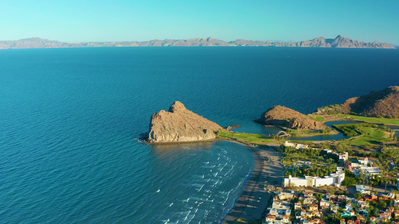 Opening aerial frame over Loreto, Baja California Sur shows a tan rocky outcrop beside blue ocean, a curving beach, shoreline buildings, distant mountains, and no visible people.