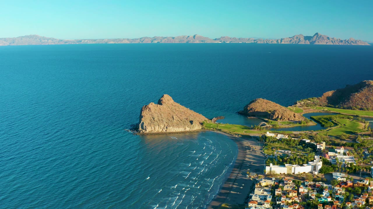 Wide aerial view of ocean, tan rock formation, curved beach, green land, and coastal buildings.