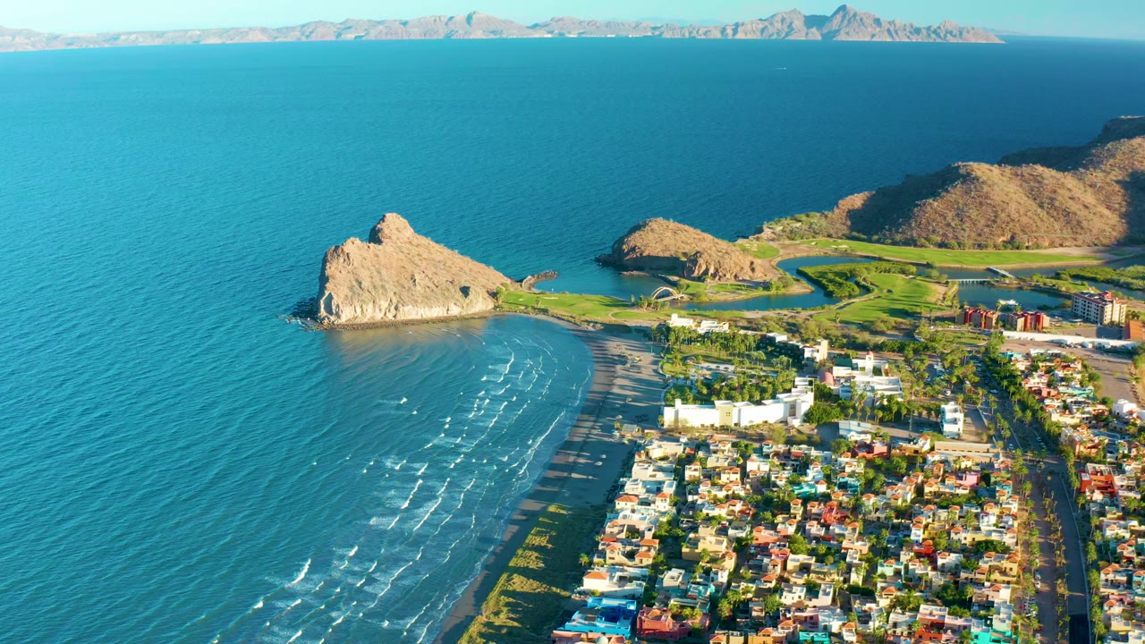 Wide aerial view over Loreto, Baja California Sur, with blue ocean at left, a curved sandy beach, a rocky outcrop near shore, residential buildings at lower right, and no visible people.