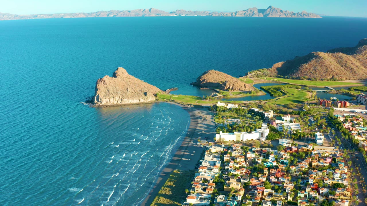 Aerial frame in Loreto, Baja California Sur, with the rocky point near center left, shallow blue water along the beach, white buildings and homes below, golf fairways behind, and no visible people.