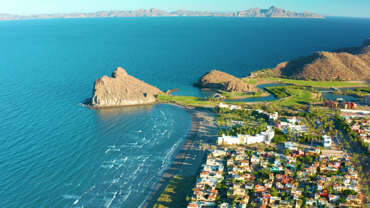 Aerial coastal scene with rocky outcrop, surf, houses, and green golf course.