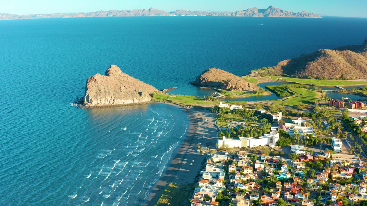 Opening aerial frame over Loreto, Baja California Sur, with turquoise water at left, a tan rock formation by the beach, town buildings at lower right, distant mountains, and no visible people.