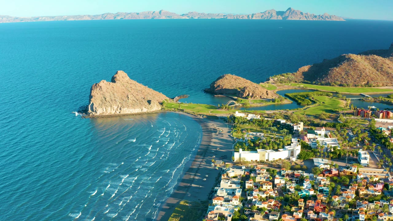 Loreto coastal aerial with broad blue water filling most of the frame, the tan headland near center left, beach surf below it, town buildings at the lower right edge, and no visible people.