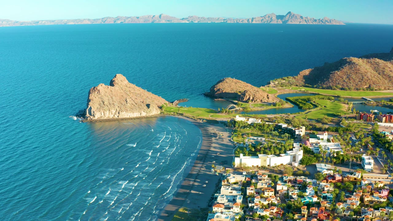 Aerial coastline view with beach, tan rock, green land, and town roofs.